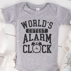 May include: A gray onesie with black text that reads "World's Cutest Alarm Clock" and a black alarm clock graphic.
