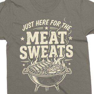 May include: A gray t-shirt with the text "JUST HERE FOR THE MEAT SWEATS" in white, above a grill graphic with ribs and a steak. The design includes stars and smoke.