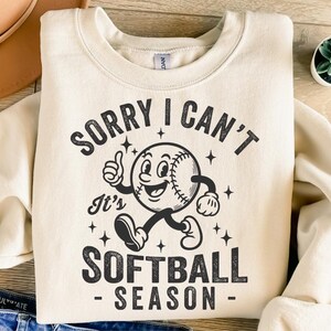 Op de afbeelding: Cr&egrave;mekleurige sweatshirt met de tekst "SORRY I CAN'T It's SOFTBALL SEASON" en een cartoon honkbalpersonage. De honkbal heeft een lachend gezicht, armen en benen. Het ontwerp is in zwart.