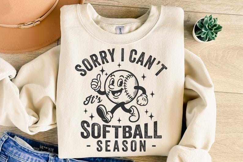 Op de afbeelding: Cr&egrave;mekleurige sweatshirt met de tekst "SORRY I CAN'T It's SOFTBALL SEASON" en een cartoon honkbalpersonage. De honkbal heeft een lachend gezicht, armen en benen. Het ontwerp is in zwart.