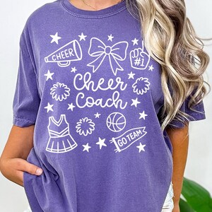 May include: Purple t-shirt with white graphic elements. The design includes a megaphone, bow, pom-poms, stars, a cheerleading uniform, a basketball, and the words "Cheer Coach" and "Go Team".