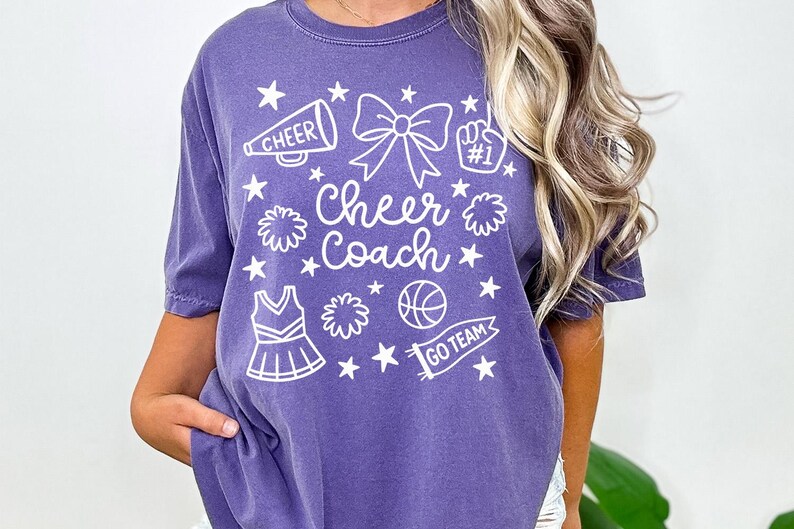 May include: Purple t-shirt with white graphic elements. The design includes a megaphone, bow, pom-poms, stars, a cheerleading uniform, a basketball, and the words "Cheer Coach" and "Go Team".