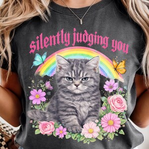 Pu&ograve; includere: T-shirt grigio scuro con un gatto tigrato grigio, arcobaleno, farfalle e fiori. Il testo "silently judging you" &egrave; presente. La maglietta ha un aspetto vintage e usurato.