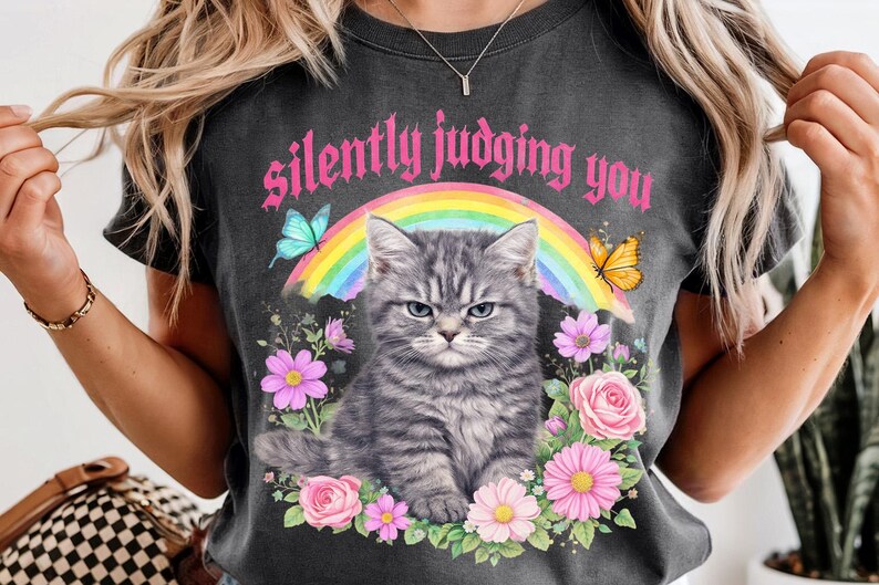 Pu&ograve; includere: T-shirt grigio scuro con un gatto tigrato grigio, arcobaleno, farfalle e fiori. Il testo "silently judging you" &egrave; presente. La maglietta ha un aspetto vintage e usurato.