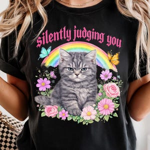 Pu&ograve; includere: T-shirt nero con un design che presenta un gatto tabby grigio con un'espressione arrabbiata, circondato da fiori e farfalle. Il testo "silently judging you" &egrave; sopra il gatto. Un arcobaleno &egrave; dietro il gatto.