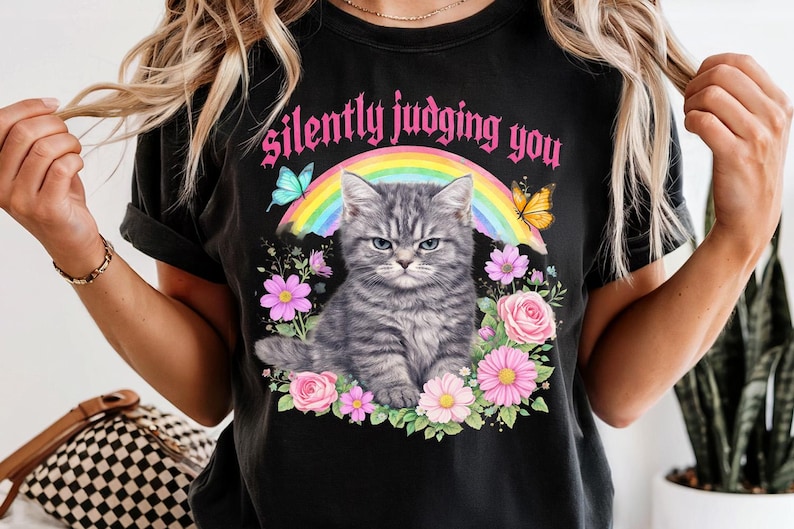 Pu&ograve; includere: T-shirt nero con un design che presenta un gatto tabby grigio con un'espressione arrabbiata, circondato da fiori e farfalle. Il testo "silently judging you" &egrave; sopra il gatto. Un arcobaleno &egrave; dietro il gatto.