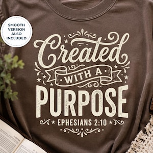 May include: A brown t-shirt with the words "Created with a Purpose" in a decorative font. The text includes the Bible verse Ephesians 2:10. A small plant, an open book, and a wooden cross are visible in the background.