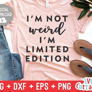 May include: A pink t-shirt with the text "I'm not weird I'm limited edition" in black lettering. The shirt is on a white background with a pair of brown sandals and a pair of blue jeans.