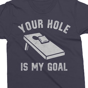 May include: Navy blue t-shirt with a white graphic of a cornhole board and the text "YOUR HOLE IS MY GOAL". The design features a bean bag on the board. A fun shirt for game day or casual wear.