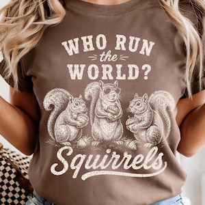 May include: A brown short-sleeved t-shirt featuring a white graphic of three squirrels, one wearing sunglasses, holding acorns. The text reads "WHO RUN the WORLD?" above the squirrels and "Squirrels" in a cursive font below the illustration.