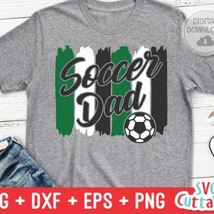May include: A gray t-shirt with a green, white, and black design that says "Soccer Dad" with a soccer ball graphic.