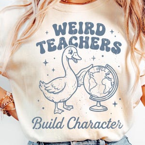 May include: Cream-colored t-shirt with a graphic featuring a goose holding a globe. The text "WEIRD TEACHERS" is arched above the goose, and "Build Character" is below. The design is in shades of blue and has a vintage, slightly distressed look.