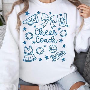 May include: White sweatshirt with a blue graphic that says "Cheer Coach." The design includes a megaphone, bow, pom-poms, a basketball, a cheerleading uniform, and stars. The phrase "Go Team" is on a banner.
