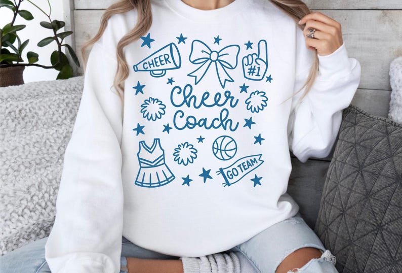 May include: White sweatshirt with a blue graphic that says "Cheer Coach." The design includes a megaphone, bow, pom-poms, a basketball, a cheerleading uniform, and stars. The phrase "Go Team" is on a banner.