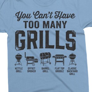 May include: Light blue t-shirt with the text "You Can't Have TOO MANY GRILLS" in a distressed font. Below the text are illustrations of different grill types: kettle, offset smoker, barrel, flat top griddle, and classic backyard grill.