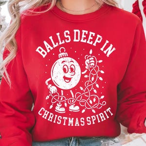 May include: Red sweatshirt with a white graphic of a smiling Christmas ornament holding string lights. The text on the sweatshirt reads "BALLS DEEP IN CHRISTMAS SPIRIT" in a distressed font. The sweatshirt is a festive holiday apparel item.