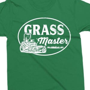 May include: A green t-shirt with a white graphic. The graphic features the words "GRASS Master" inside an oval, with a lawn mower illustration below the text. The shirt is made of a soft material.