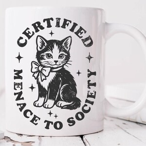 May include: White ceramic mug featuring a black and white illustration of a cat wearing a bow tie, surrounded by the text "CERTIFIED MENACE TO SOCIETY" in a circular design. The mug has a curved handle.