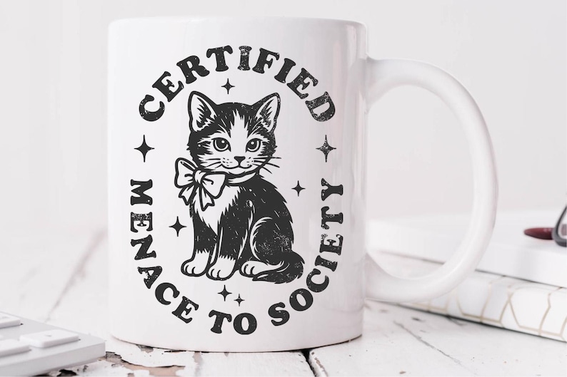 May include: White ceramic mug featuring a black and white illustration of a cat wearing a bow tie, surrounded by the text "CERTIFIED MENACE TO SOCIETY" in a circular design. The mug has a curved handle.