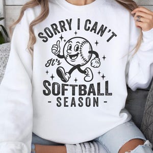 Op de afbeelding: Witte sweatshirt met een zwart-wit softbalafbeelding. De afbeelding toont een softbalpersonage met de tekst "SORRY I CAN'T It's SOFTBALL SEASON". Het ontwerp bevat sterren en een duim omhoog.