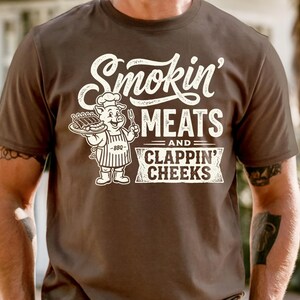 May include: Brown t-shirt with the text "Smokin' MEATS and CLAPPIN' CHEEKS" in white. A cartoon pig wearing a chef's hat and apron is holding a platter of ribs and a fork.