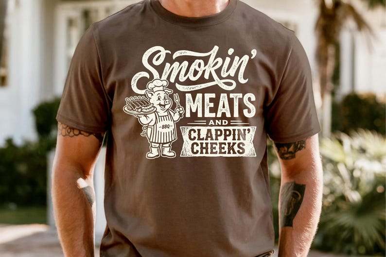 May include: Brown t-shirt with the text "Smokin' MEATS and CLAPPIN' CHEEKS" in white. A cartoon pig wearing a chef's hat and apron is holding a platter of ribs and a fork.