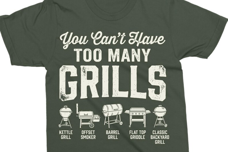 May include: Dark green t-shirt with the text "You Can't Have TOO MANY GRILLS" in white. Below the text are illustrations of different grill types: kettle, offset smoker, barrel, flat top griddle, and classic backyard grill.