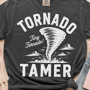 May include: A charcoal gray short-sleeve t-shirt featuring a white graphic print of a tornado. The text reads TORNADO TAMER in large block letters with Tiny Tornado! written in script. The design includes small clouds and debris at the base.