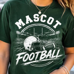 May include: A dark green short-sleeve t-shirt featuring a white graphic print of a football stadium, a helmet, and a football. The text MASCOT is printed in a bold white font above the stadium, and the word FOOTBALL is printed below in white.