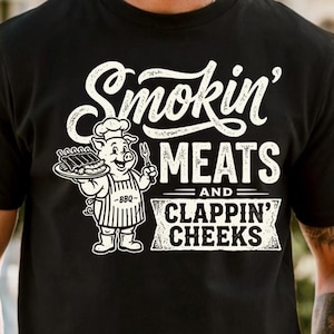 May include: Black t-shirt with a white graphic. The graphic features the text "Smokin' Meats and Clappin' Cheeks" in a stylized font. A cartoon pig wearing a chef's hat and apron is holding a plate of ribs and a fork.