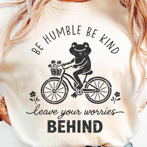 May include: Cream-coloured t-shirt featuring a black graphic of a frog on a bicycle, with flowers in a basket. The text says "BE HUMBLE BE KIND" above the frog and "leave your worries BEHIND" below.
