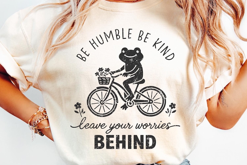 May include: Cream-coloured t-shirt featuring a black graphic of a frog on a bicycle, with flowers in a basket. The text says "BE HUMBLE BE KIND" above the frog and "leave your worries BEHIND" below.