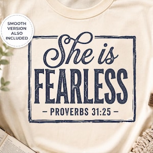 May include: Cream-colored t-shirt with the text "She is Fearless - Proverbs 31:25" in a navy blue, distressed font. A small, white circle with text is in the upper left corner. A wooden cross and open book are in the background.