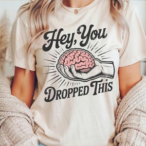 May include: Beige t-shirt with the text "Hey, You Dropped This" in a vintage style. The graphic features a pink brain held in a hand. The shirt is a casual, everyday wear item.
