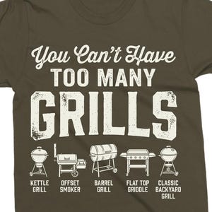 May include: Olive green t-shirt with the text "You Can't Have TOO MANY GRILLS" in white. Below the text are illustrations of different grill types, including kettle, offset smoker, barrel, flat top griddle, and classic backyard grills.