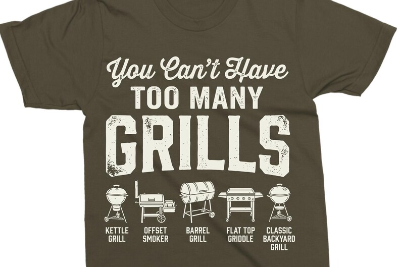 May include: Olive green t-shirt with the text "You Can't Have TOO MANY GRILLS" in white. Below the text are illustrations of different grill types, including kettle, offset smoker, barrel, flat top griddle, and classic backyard grills.