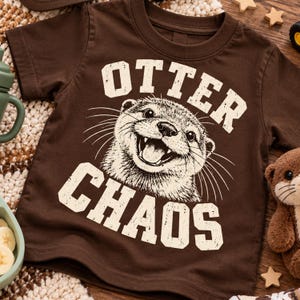 May include: A dark brown short-sleeve t-shirt featuring a graphic print of a smiling otter face in the centre. The text "OTTER" is printed in a distressed cream font above the otter, and the word "CHAOS" is printed in the same font below.
