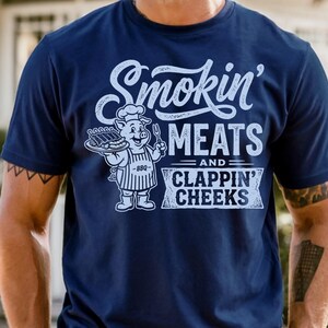 May include: Navy blue t-shirt with the text "Smokin' Meats and Clappin' Cheeks" in white script. A cartoon pig wearing a chef's hat and apron is holding a tray of ribs and a fork. The shirt has short sleeves.