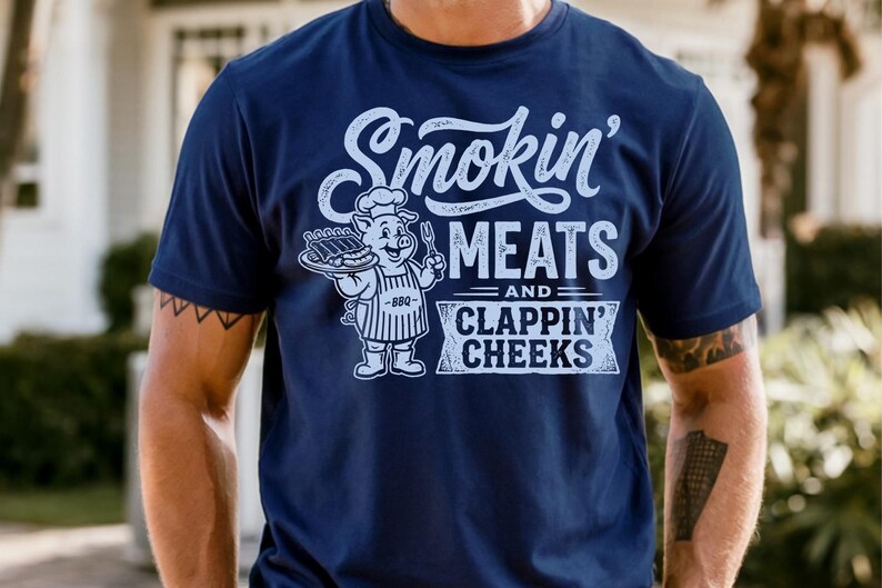 May include: Navy blue t-shirt with the text "Smokin' Meats and Clappin' Cheeks" in white script. A cartoon pig wearing a chef's hat and apron is holding a tray of ribs and a fork. The shirt has short sleeves.