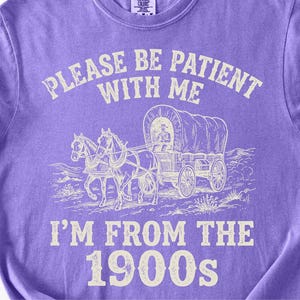 May include: A purple t-shirt with the text "PLEASE BE PATIENT WITH ME" above an illustration of a covered wagon pulled by two horses. Below the illustration is the text "I'M FROM THE 1900s".