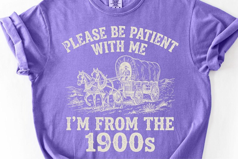 May include: A purple t-shirt with the text "PLEASE BE PATIENT WITH ME" above an illustration of a covered wagon pulled by two horses. Below the illustration is the text "I'M FROM THE 1900s".