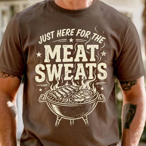 May include: Brown t-shirt with the text "JUST HERE FOR THE MEAT SWEATS" in a distressed font. The graphic features a grill with ribs and a steak. The shirt is short-sleeved.