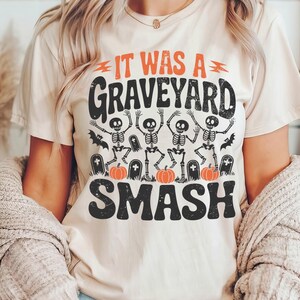 May include: Beige t-shirt with the text "IT WAS A GRAVEYARD SMASH" in black and orange. The graphic features dancing skeletons, bats, pumpkins, and tombstones, perfect for Halloween.