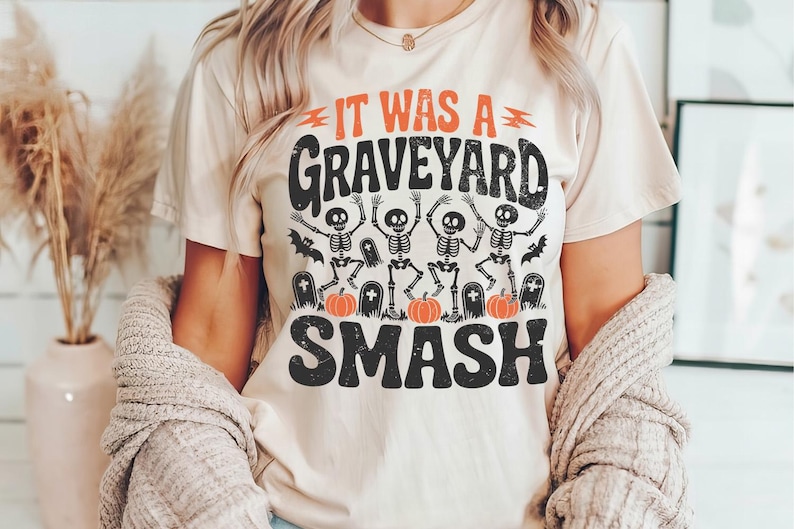 May include: Beige t-shirt with the text "IT WAS A GRAVEYARD SMASH" in black and orange. The graphic features dancing skeletons, bats, pumpkins, and tombstones, perfect for Halloween.
