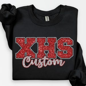 May include: Black sweatshirt with the text "XHS Custom" in red glitter letters with a silver outline. The sweatshirt is a crew neck style and has long sleeves. The Gildan brand tag is visible.