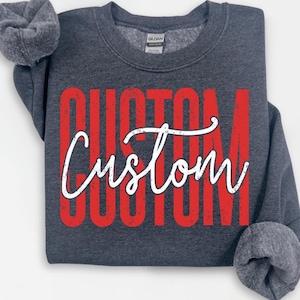 以下が含まれることがあります： 大きな赤いブロック文字で「CUSTOM」という文字と、白い筆記体の「Custom」が重ねられた濃い灰色のスウェットシャツ。スウェットシャツはクルーネックで、グレーの袖口が付いた長袖です。