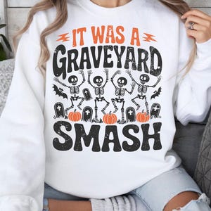 May include: White sweatshirt with a Halloween-themed graphic. The design features the text "IT WAS A GRAVEYARD SMASH" in black and orange, along with illustrations of skeletons, pumpkins, and tombstones. A fun and festive design for the spooky season.