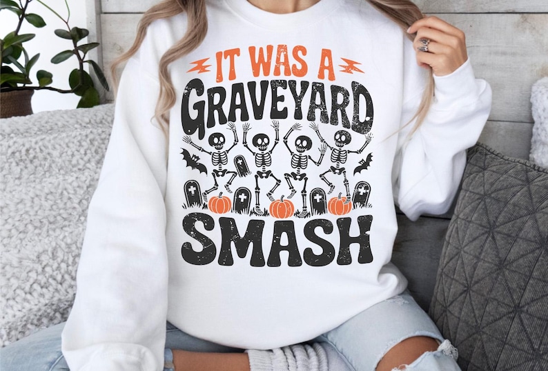 May include: White sweatshirt with a Halloween-themed graphic. The design features the text "IT WAS A GRAVEYARD SMASH" in black and orange, along with illustrations of skeletons, pumpkins, and tombstones. A fun and festive design for the spooky season.