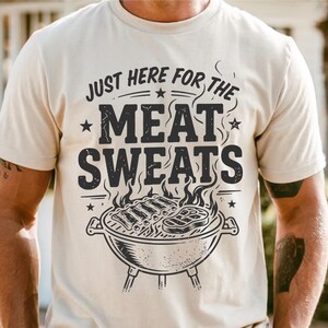 May include: Cream-colored t-shirt with a graphic design. The design features the text "JUST HERE FOR THE MEAT SWEATS" above a charcoal grill with ribs and a steak. The shirt is short-sleeved.