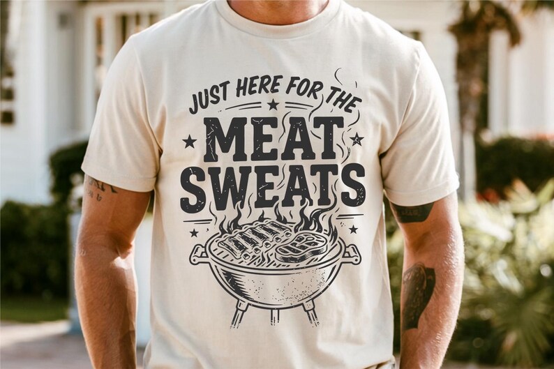 May include: Cream-colored t-shirt with a graphic design. The design features the text "JUST HERE FOR THE MEAT SWEATS" above a charcoal grill with ribs and a steak. The shirt is short-sleeved.